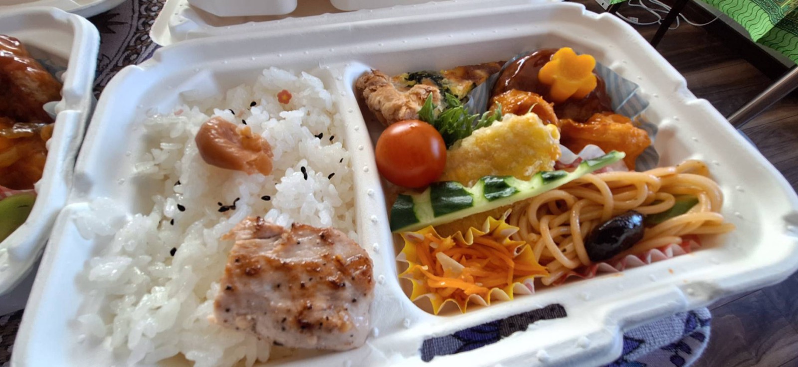 お弁当のアップ。ご飯、鶏の照り焼き、キッシュ、キャロットラペなどが彩りよく詰まっている